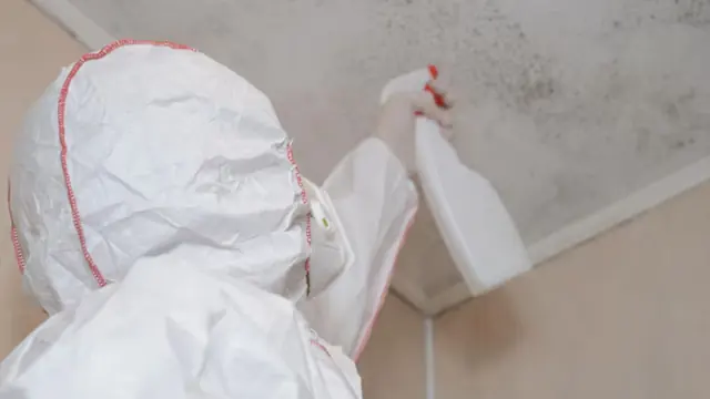 Person in a hooded suit spraying a ceiling corner to treat mold
